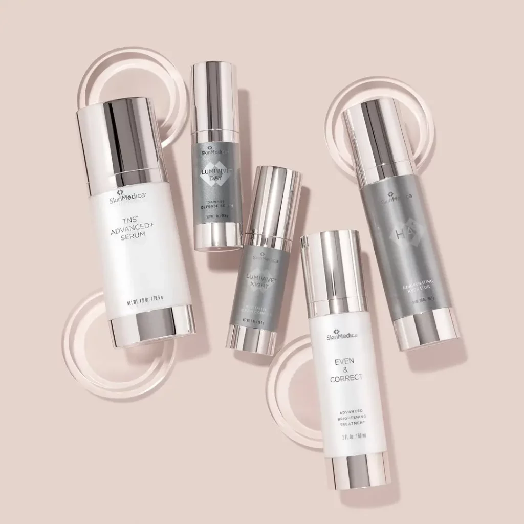 A collection of five SkinMedica skincare products arranged aesthetically on a soft beige background. The products include TNS Advanced+ Serum, two Lumivive Day Defense and Night Revitalize serums, HA5 Rejuvenating Hydrator, and Even Correct Advanced Pigment Correcting Cream, all in sleek, metallic dispensers with the brand logo visible.