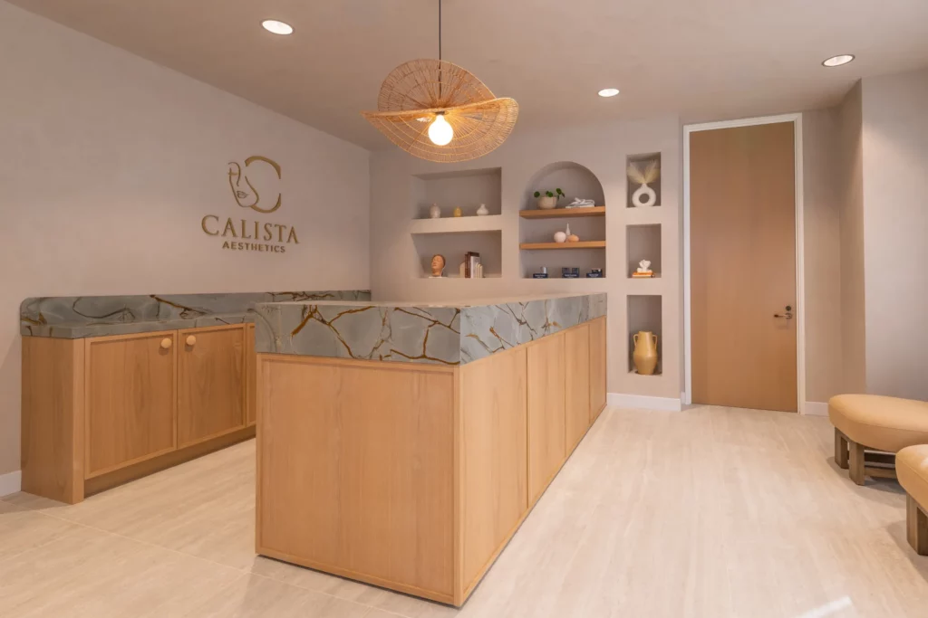 Reception area of Calista Aesthetics with a marble-accented wooden counter, decorative shelving, and minimalist decor under soft lighting.