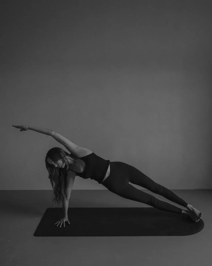 Black-and-white photo of a woman doing a Pilates side plank on a mat, top arm reaching overhead, feet stacked, in a minimal studio setting