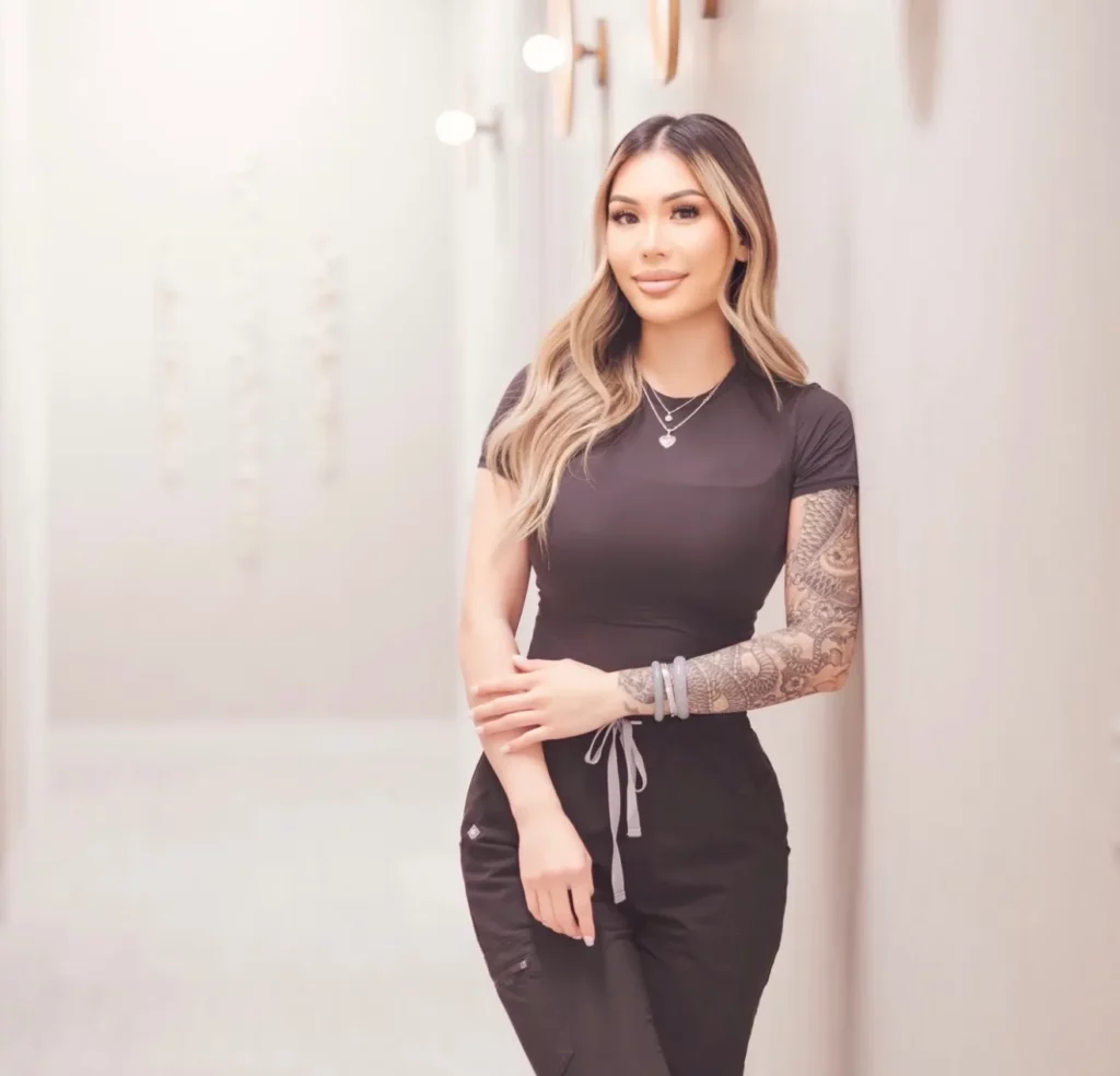 Portrait of a woman with long highlighted hair wearing a black outfit, showing a detailed sleeve tattoo, standing against a soft neutral background