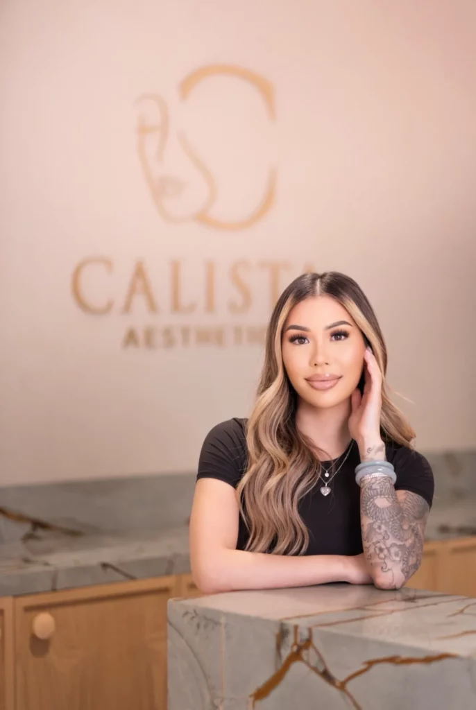 Woman in black outfit standing in a softly lit hallway with modern wall sconces Woman sitting on a cushioned chair in a modern lounge with indoor olive tree, wearing black outfit and white sneakers Woman standing with arms crossed in a bright hallway with arched doorway and minimalist decor Woman leaning on a marble reception desk with clinic logo in background, wearing black outfit and showing arm tattoo